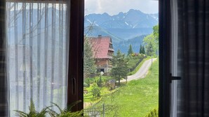 Vista per una finestra amb cortines lleugeres, que mostra un paisatge rural amb una casa, un camí sinuós i muntanyes al fons.