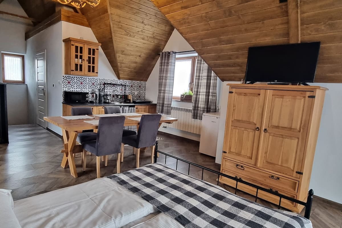 A cozy loft-style room with a dining area, a sofa, a TV, and a kitchen in the background.