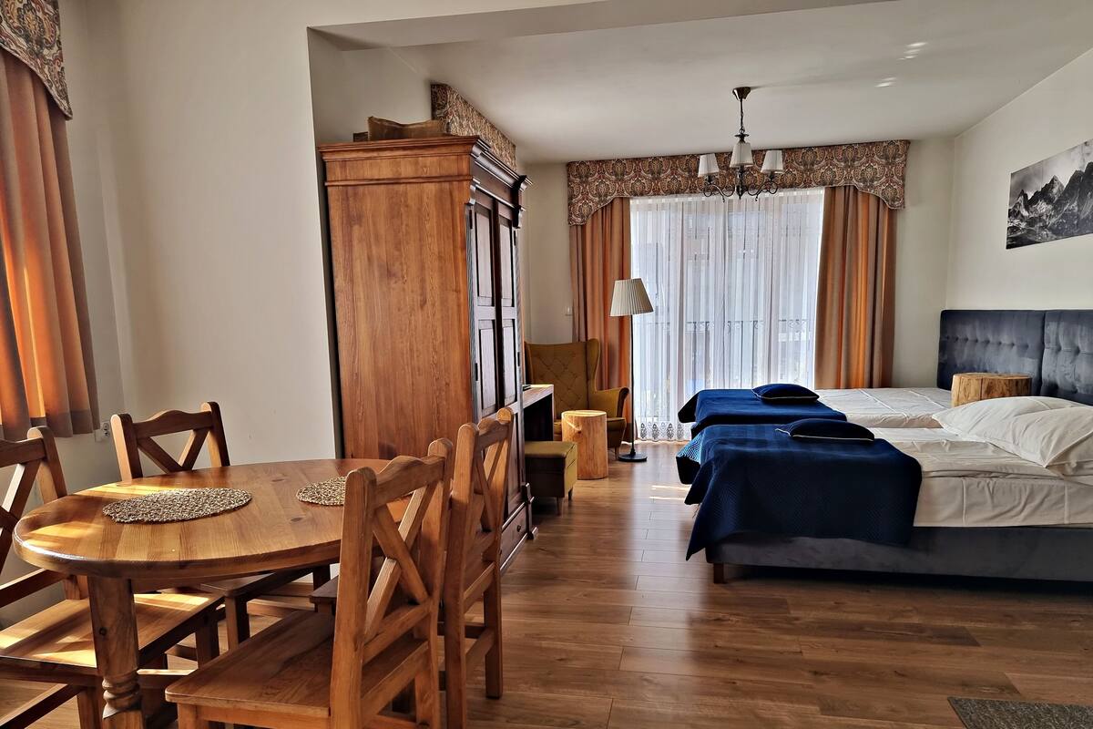 A hotel room with a wooden dining table, chairs, a cabinet, a bed with two beds, a lamp, and a window with curtains.