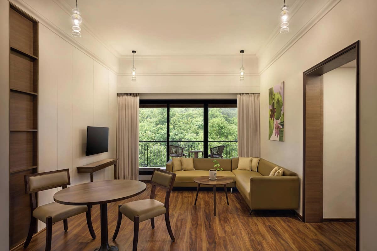 Una sala de estar moderna con un sofá, una mesa de comedor y sillas. Hay un televisor montado en la pared, una ventana con cortinas y un balcón con vistas a vegetación.