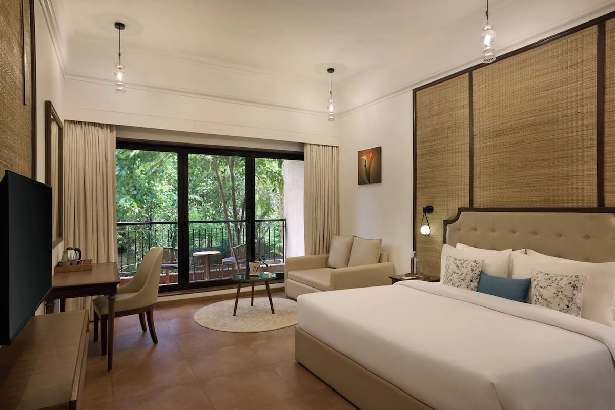 Habitación de hotel con una cama grande, un escritorio con silla, un sofá, una mesa de comedor y vistas a zonas verdes a través de las ventanas.