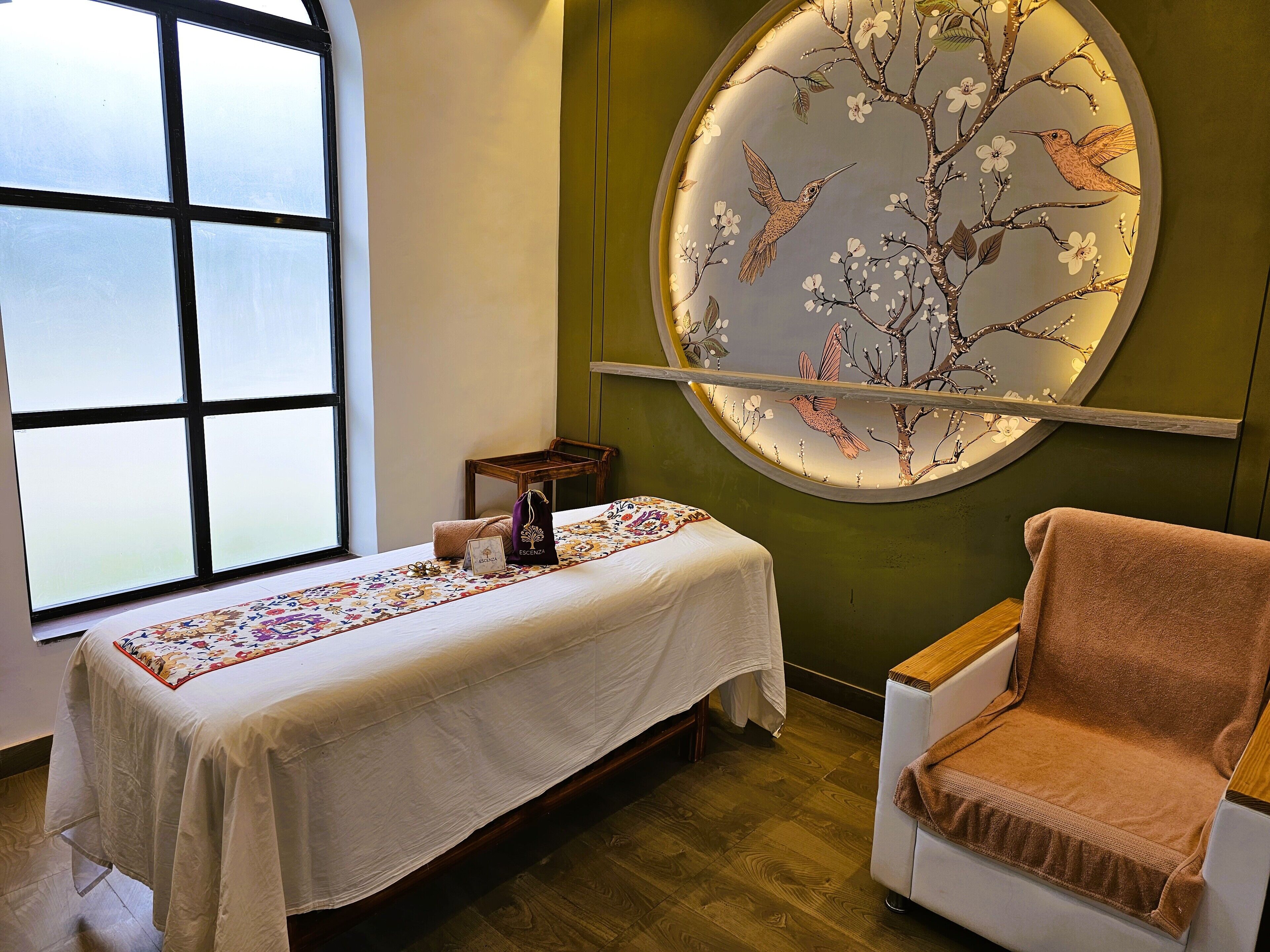 Ein Zimmer mit einem Bett, einem Sessel, einem Tisch mit Tischdecke, einem Fenster und einem Wandbild.