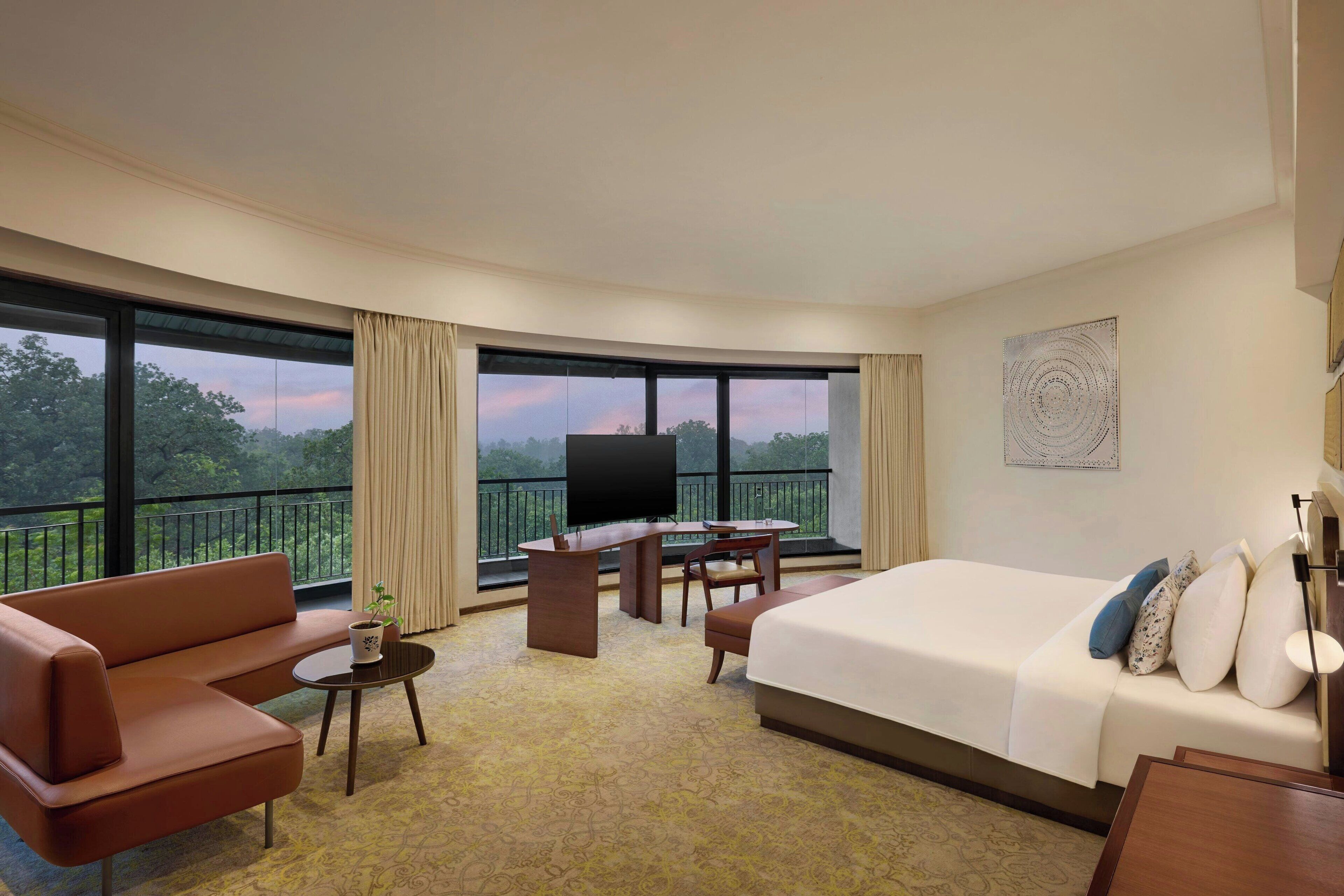 Habitación de hotel con una cama grande, un sofá, un escritorio y un televisor. Hay un balcón con vistas a árboles y un atardecer.