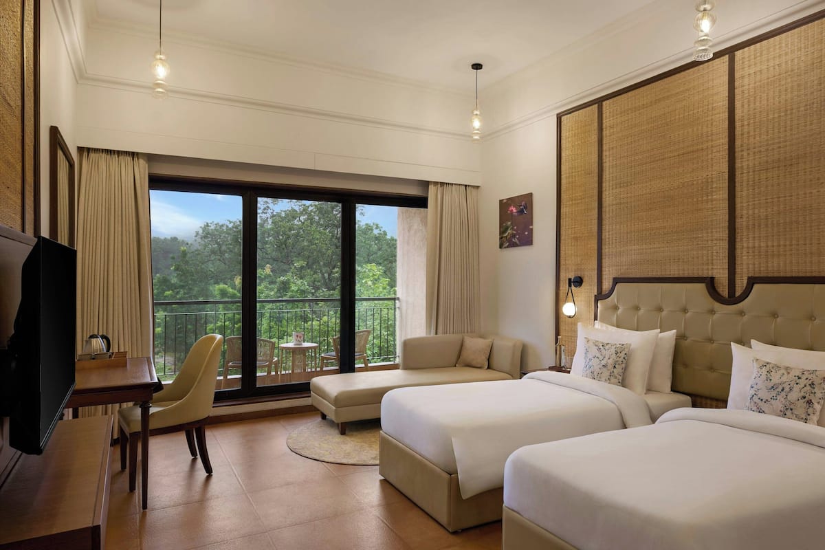 Habitación de hotel con dos camas, televisión, balcón con mesa y sillas, y vista a árboles.