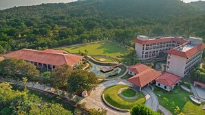 A large building with multiple red-tiled roofs, surrounded by lush greenery and a well-maintained garden with a circular water feature.