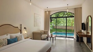 A hotel room with a large bed, a sofa, a small table, a TV, and a view of a swimming pool and greenery.