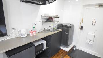 Cozinha compacta com micro-ondas, pia e pequena geladeira. Há prateleiras sobre o balcão com xícaras e uma chaleira.
