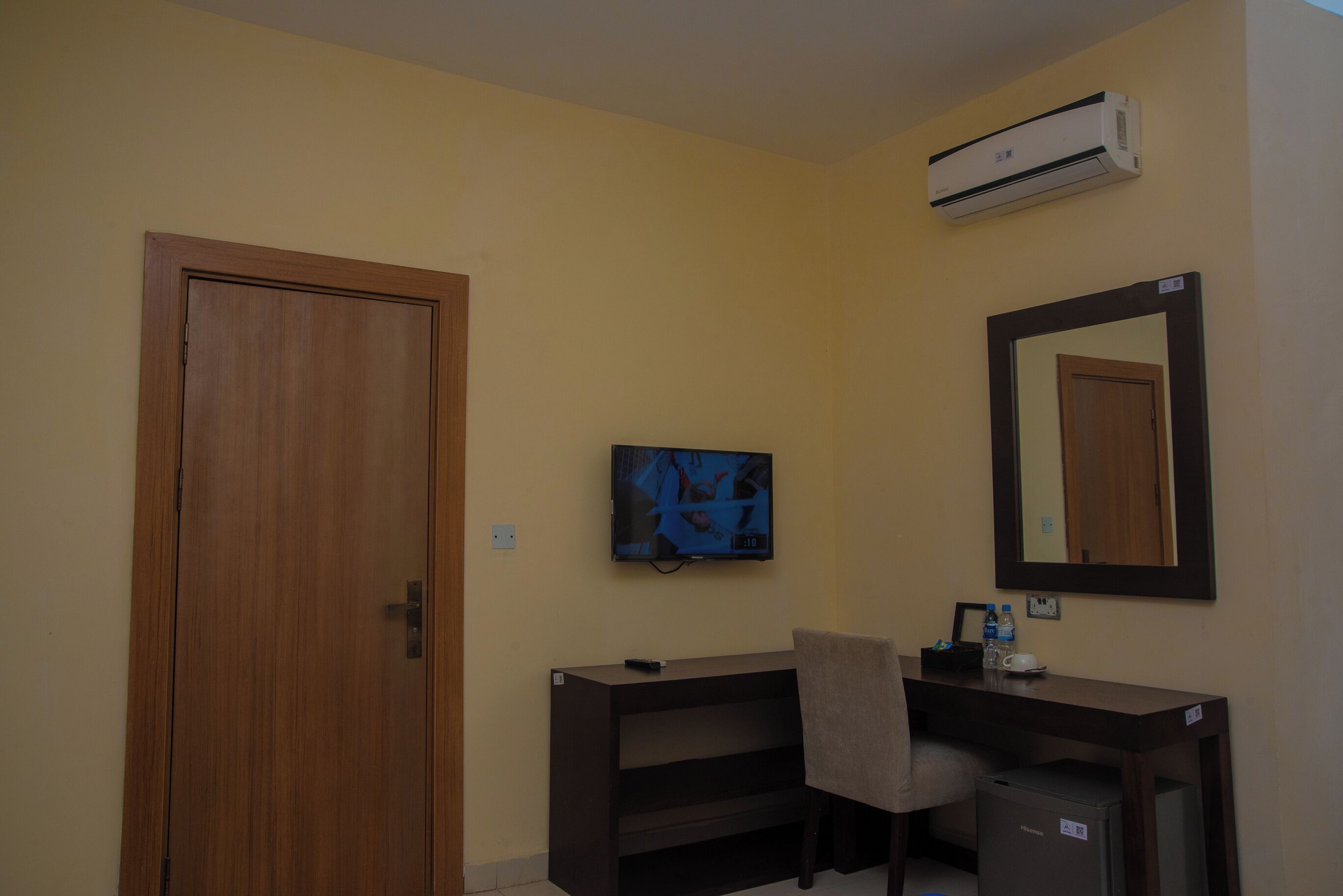 Habitación de hotel con puerta de madera, un escritorio con silla, televisor, espejo y equipo de aire acondicionado.