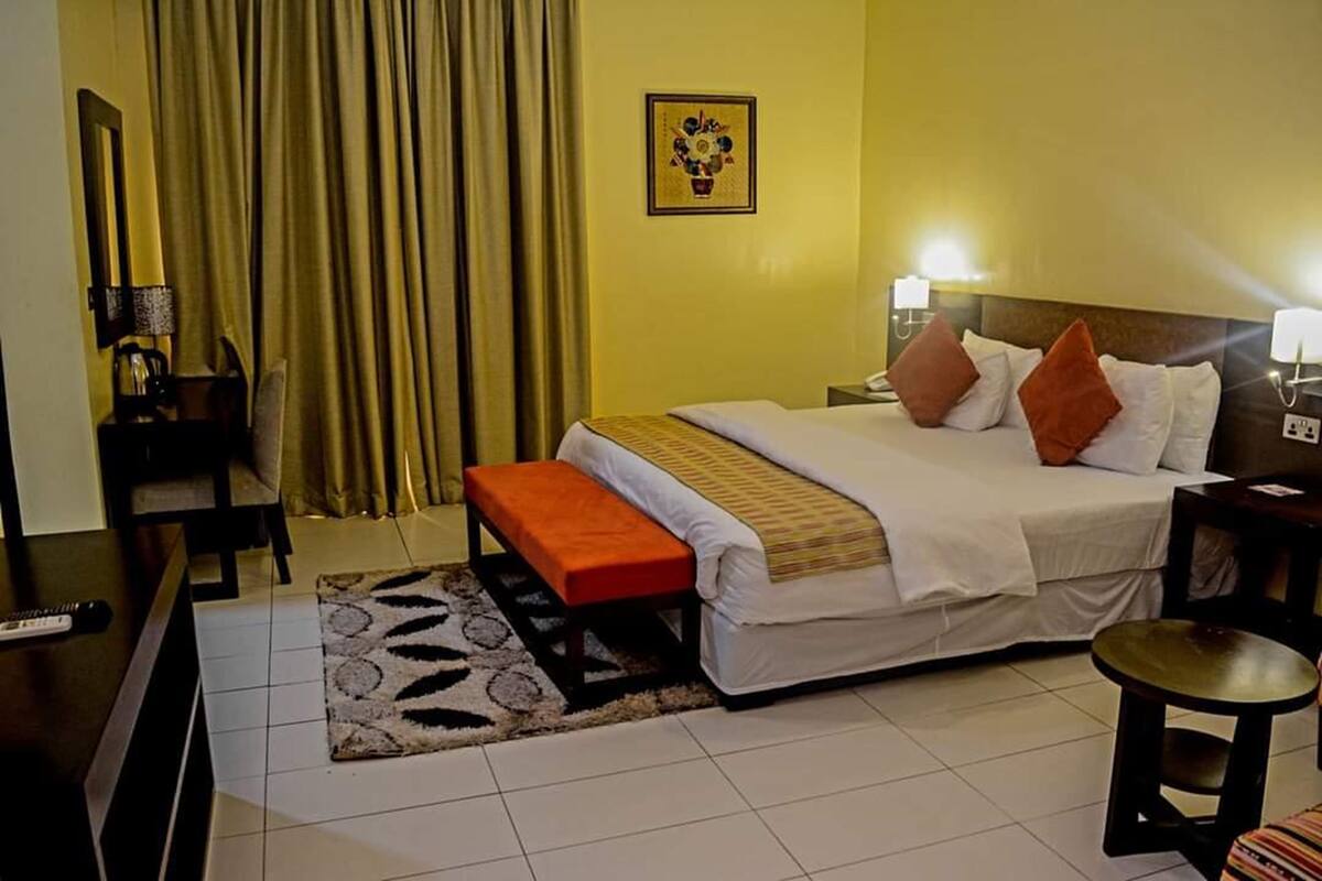 A hotel room with a bed, a bench, a small table, a lamp, and a picture on the wall.