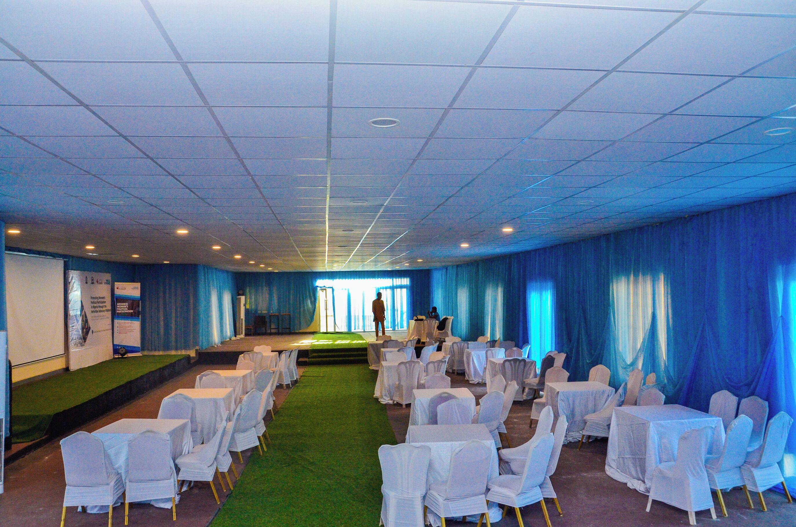 A banquet hall with rows of tables and chairs, a stage, and blue curtains.