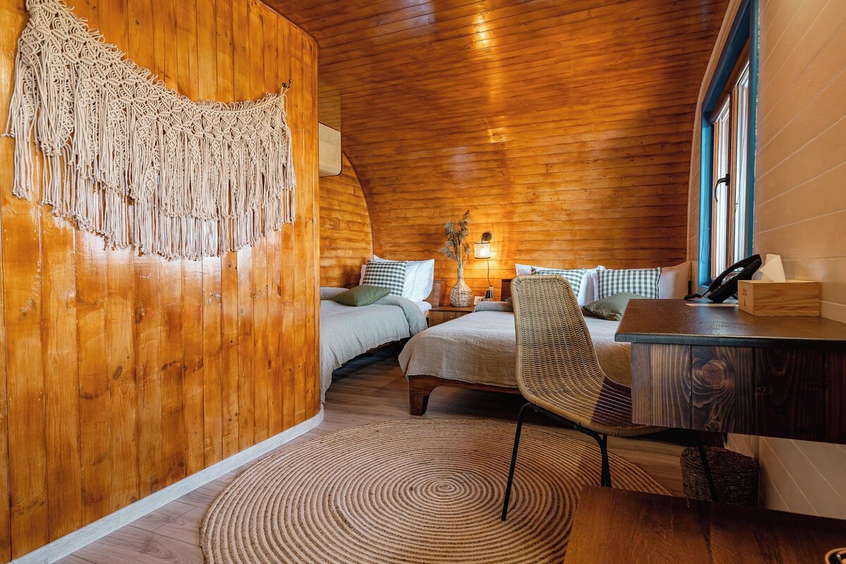 A cozy room with wooden walls, a bed with checkered bedding, a wicker chair, a desk with a computer, and a macramé wall hanging.