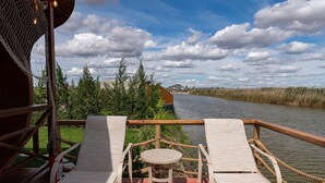 A wooden deck with two lounge chairs and a small table, overlooking a river with a house in the background. - Razelm Luxury Resort (Jurilovca)