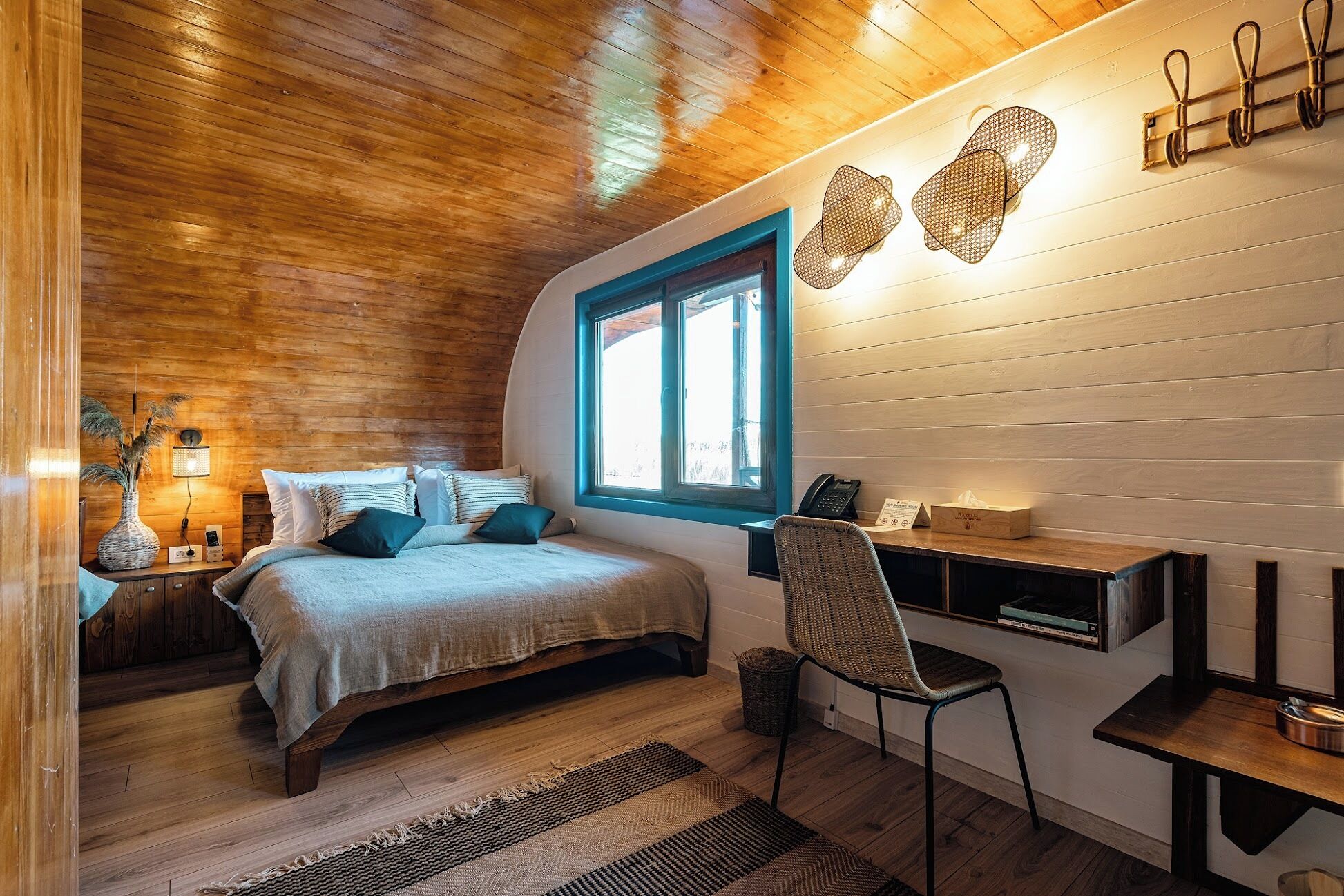 Un dormitorio acogedor con techo de madera, una cama con marco de ventana azul, un escritorio con silla y una mesita de noche con lámpara.