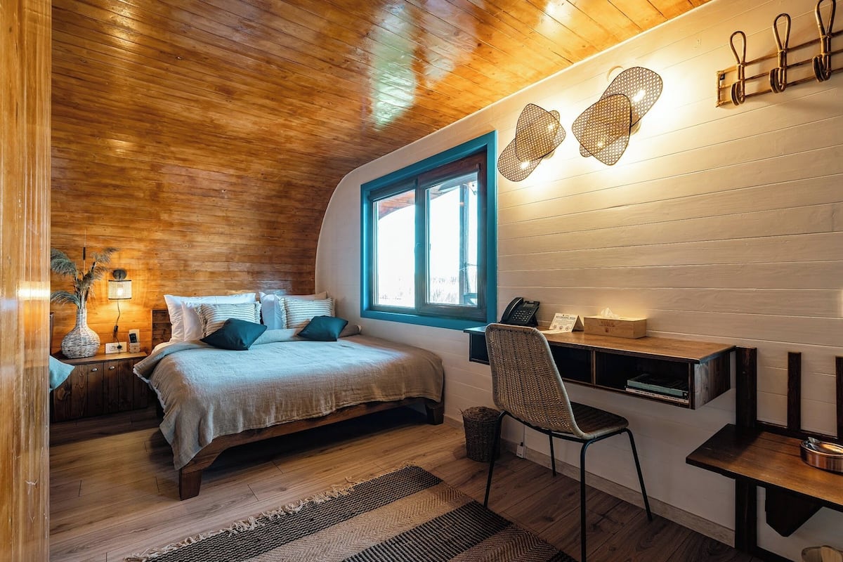 A cozy bedroom with a wooden ceiling, a bed with a blue window frame, a desk with a chair, and a bedside table with a lamp.