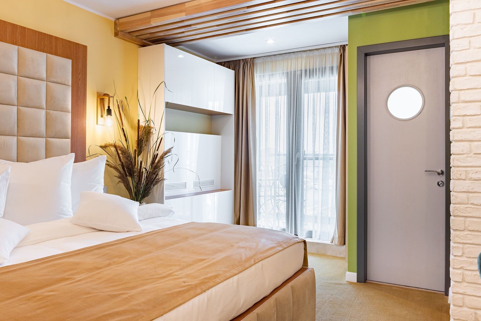 Una habitación de hotel moderna con una cama grande, cabecera de madera, armario empotrado, espejo redondo y vistas al exterior a través de un amplio ventanal con cortinas translúcidas.