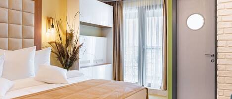 Una habitación de hotel moderna con una cama grande, cabecera de madera, armario empotrado, espejo redondo y vistas al exterior a través de un amplio ventanal con cortinas translúcidas.