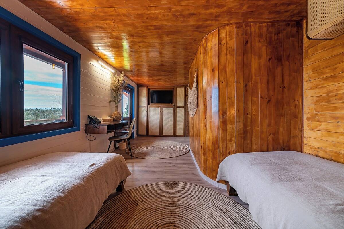 A cabin-style room with two beds, a desk with a chair, a window with a view, and a wooden ceiling.
