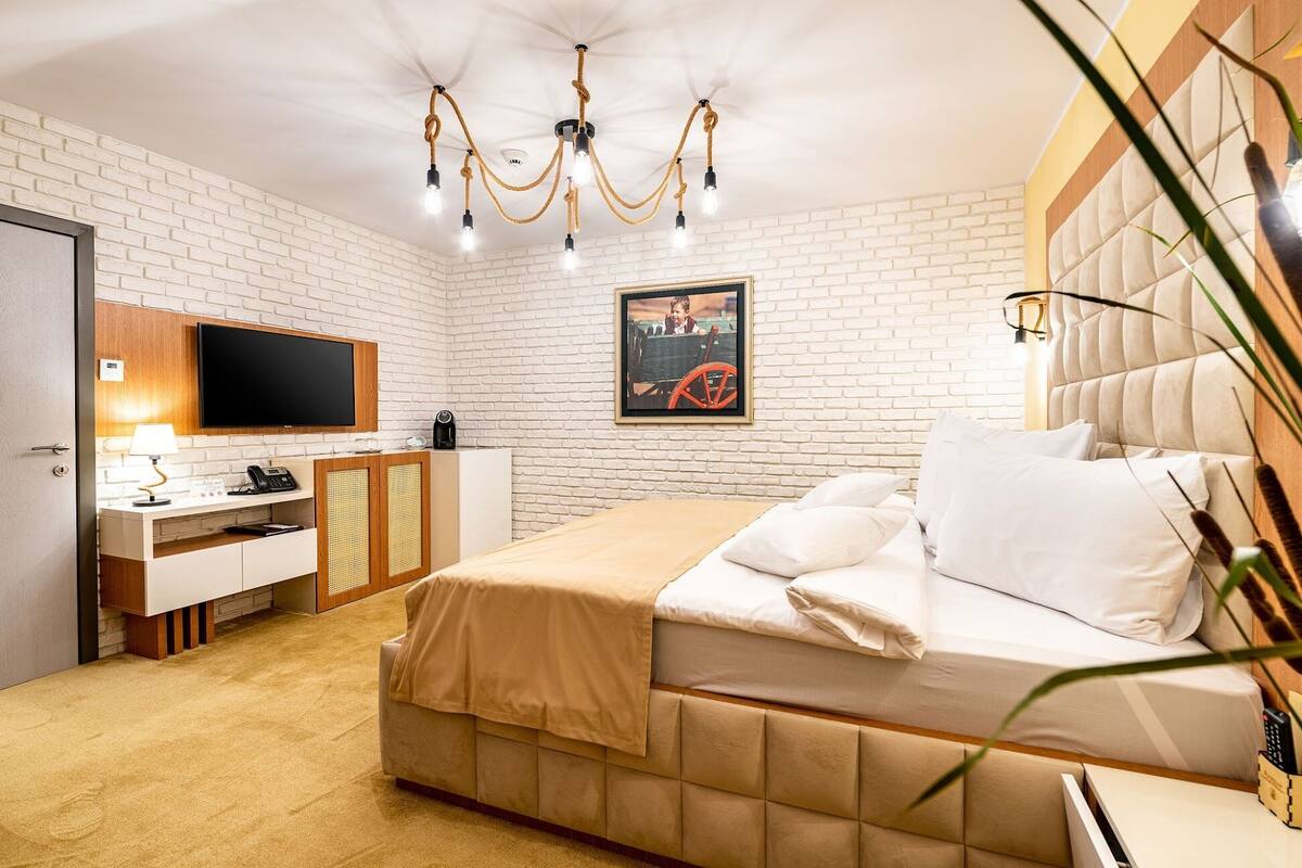A hotel room with a white brick wall, a bed with white bedding, a desk with a computer and lamp, a television, and a chandelier.
