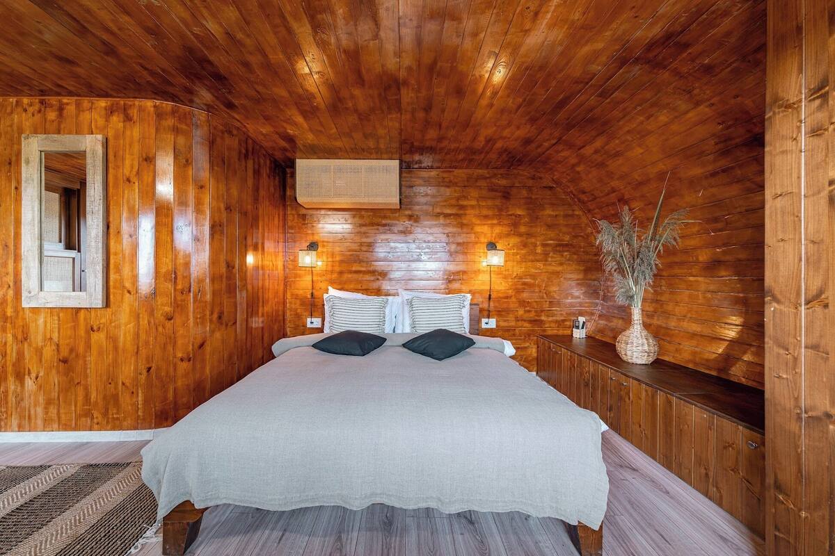 A wooden-paneled room with a bed, two wall-mounted lights, a woven basket, and a vase with dried plants.