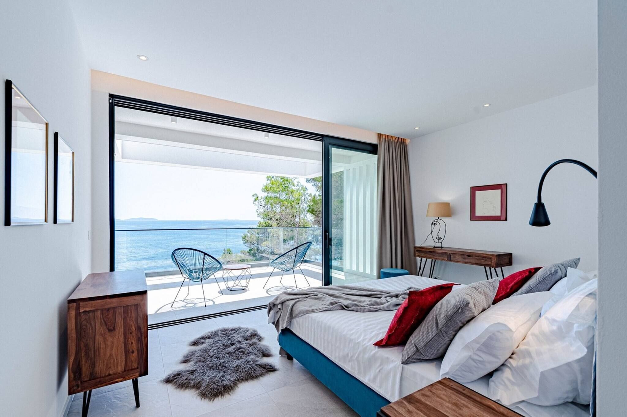 A modern bedroom with a large bed, a view of the sea, a balcony with chairs, and a wooden side table.