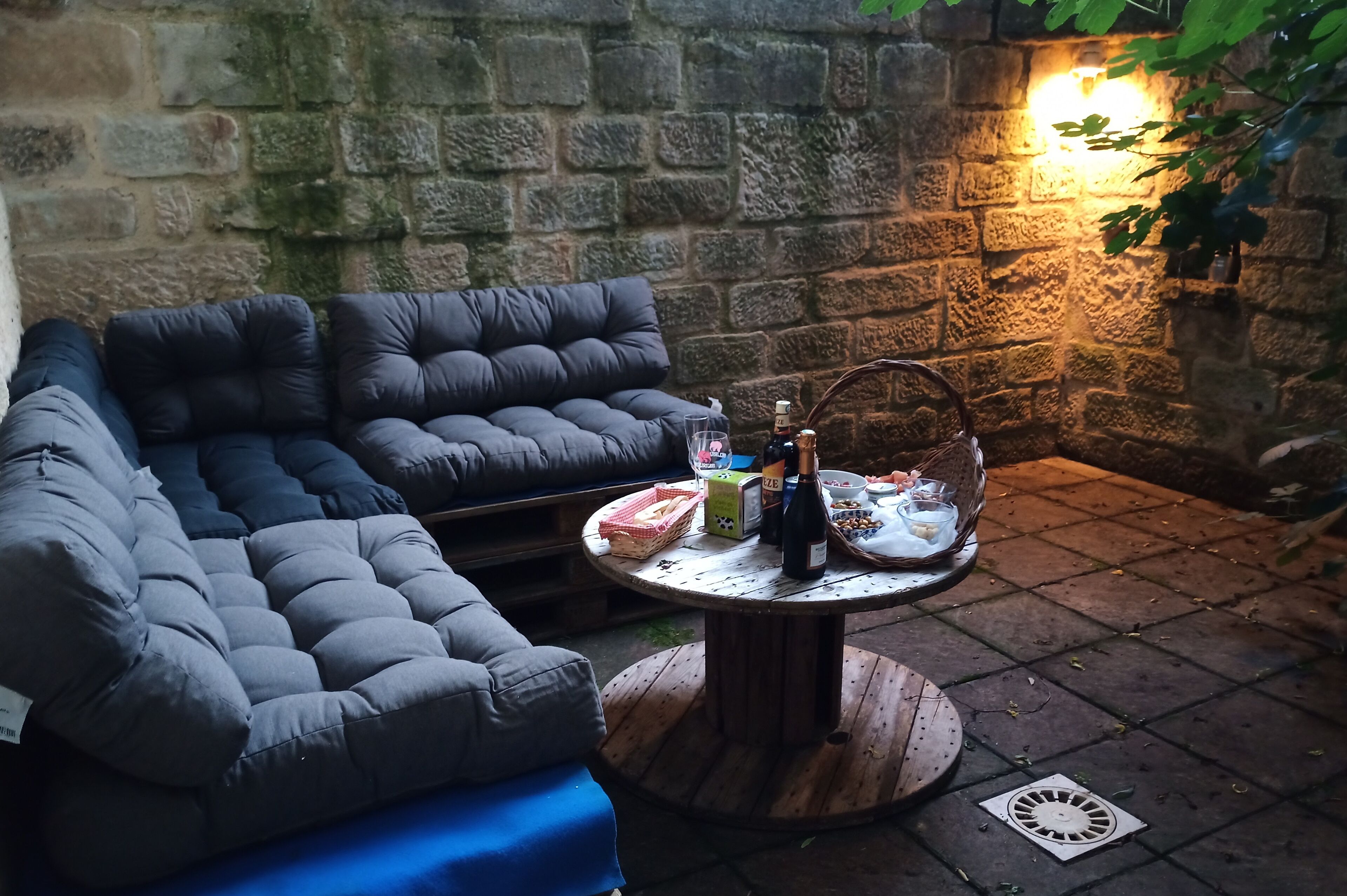 A cozy outdoor seating area with a cushioned sofa and a round table with bottles and snacks.