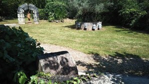 A stone archway and a stone structure with a circular base, surrounded by grass and trees.