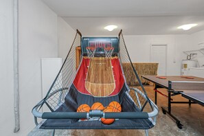 Uma tabela de basquete com placar digital, uma mesa de pingue-pongue e outra mesa de pingue-pongue em um espaço com paredes brancas e piso de madeira.