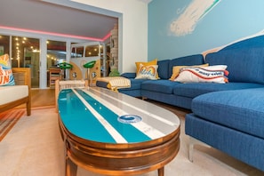 Uma sala de estar com um sofá azul, uma mesa de centro com o desenho de uma prancha de surf e uma parede com um mural tropical.