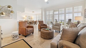 Ein geräumiges Wohnzimmer mit Deckenventilator, großen Fenstern mit Jalousien und einem Essbereich im Hintergrund.