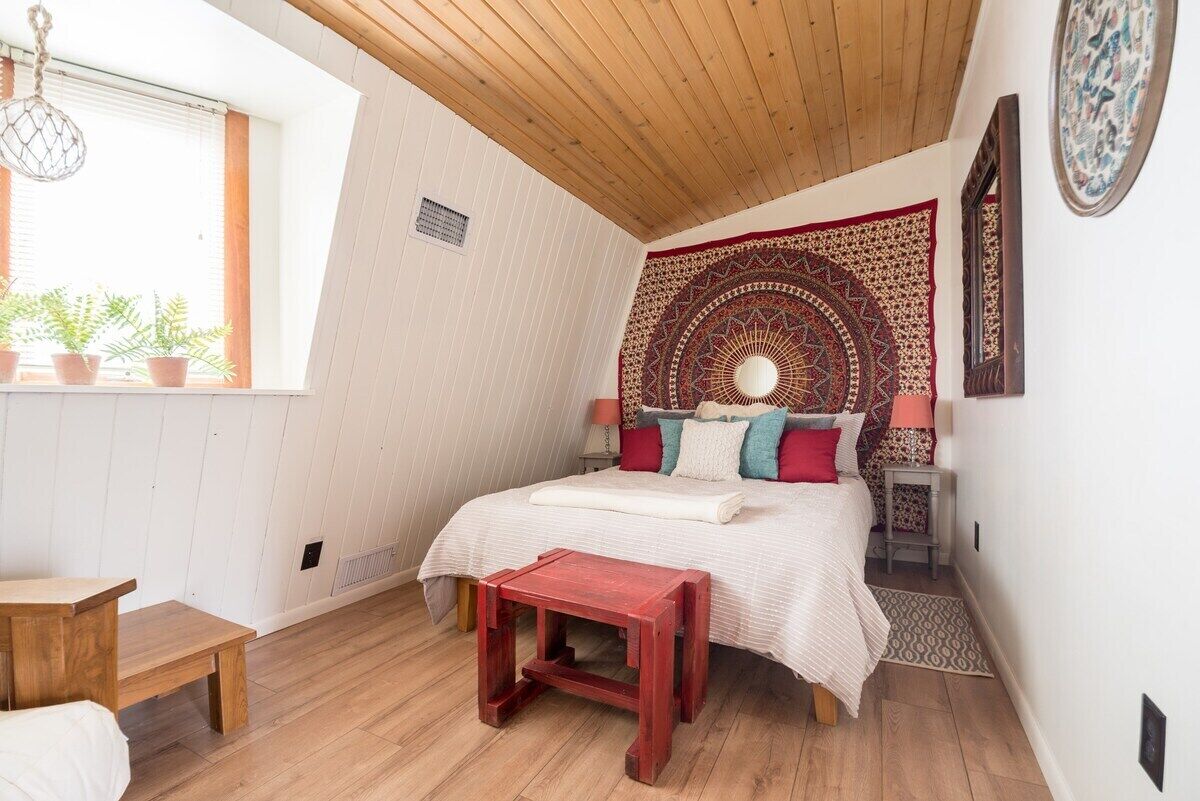 A cozy bedroom with a wooden ceiling, a large bed with colorful pillows, a red bench, and a window with plants.