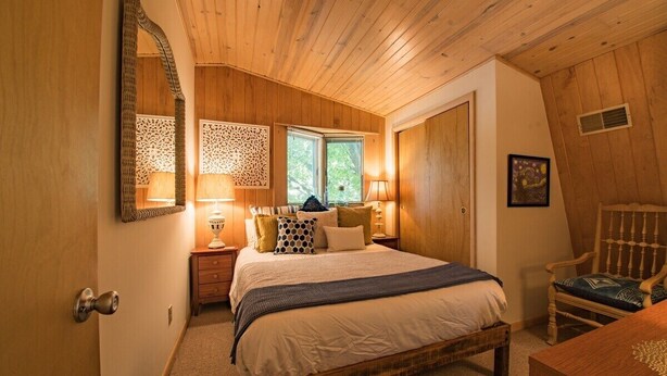 Un dormitorio con techo de madera, una cama grande con muchas almohadas, una mesita de noche con lámpara y una silla.