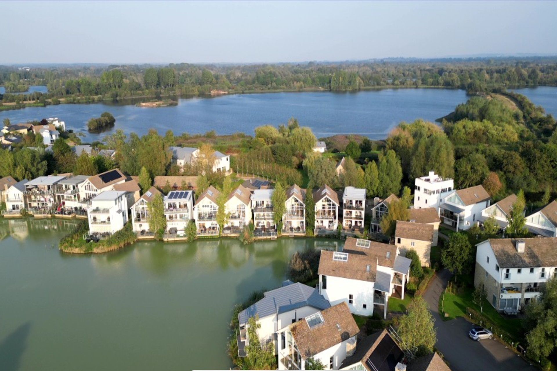 A lakeside residential area with houses featuring balconies and solar panels, surrounded by dense greenery and a calm lake.
