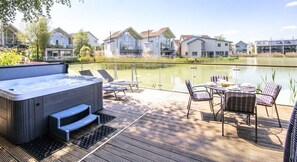 Een hottub op een houten terras, met stoelen en een tafel, met uitzicht op een rustig wateroppervlak en moderne huizen.