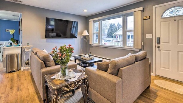 A living room with a sofa, a coffee table, a TV mounted on the wall, and a window with a view of houses outside. - Homelike, peaceful and centrally- home located in the hamlet of Rocky  Point  NY (Rocky Point)