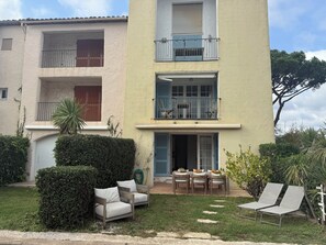 Un bâtiment à deux étages doté d’un balcon, d’un petit jardin avec une table et des chaises, et d’une haie.