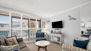 A living room with a sofa, armchair, and a round coffee table. There is a TV mounted on the wall, a lamp on a side table, and a window with blinds. - Ocean Block 2 BR 2 BA Unit - Short Walk to the Beach! (Ocean City)