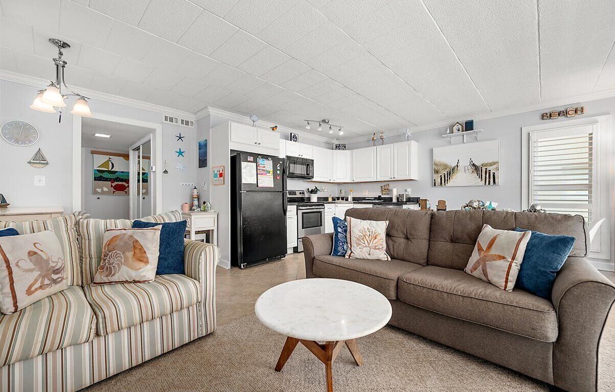 A spacious living room with a striped sofa, a round coffee table, and a brown sofa with decorative pillows. A kitchen with white cabinets and a black refrigerator is visible in the background.