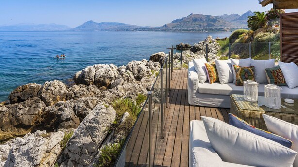 A wooden deck with white outdoor furniture overlooking a calm sea and distant mountains. - Rais house with private access to the sea (Solanto)