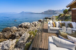 A wooden deck with white outdoor furniture overlooking a calm sea and distant mountains. - Rais house with private access to the sea (Solanto)