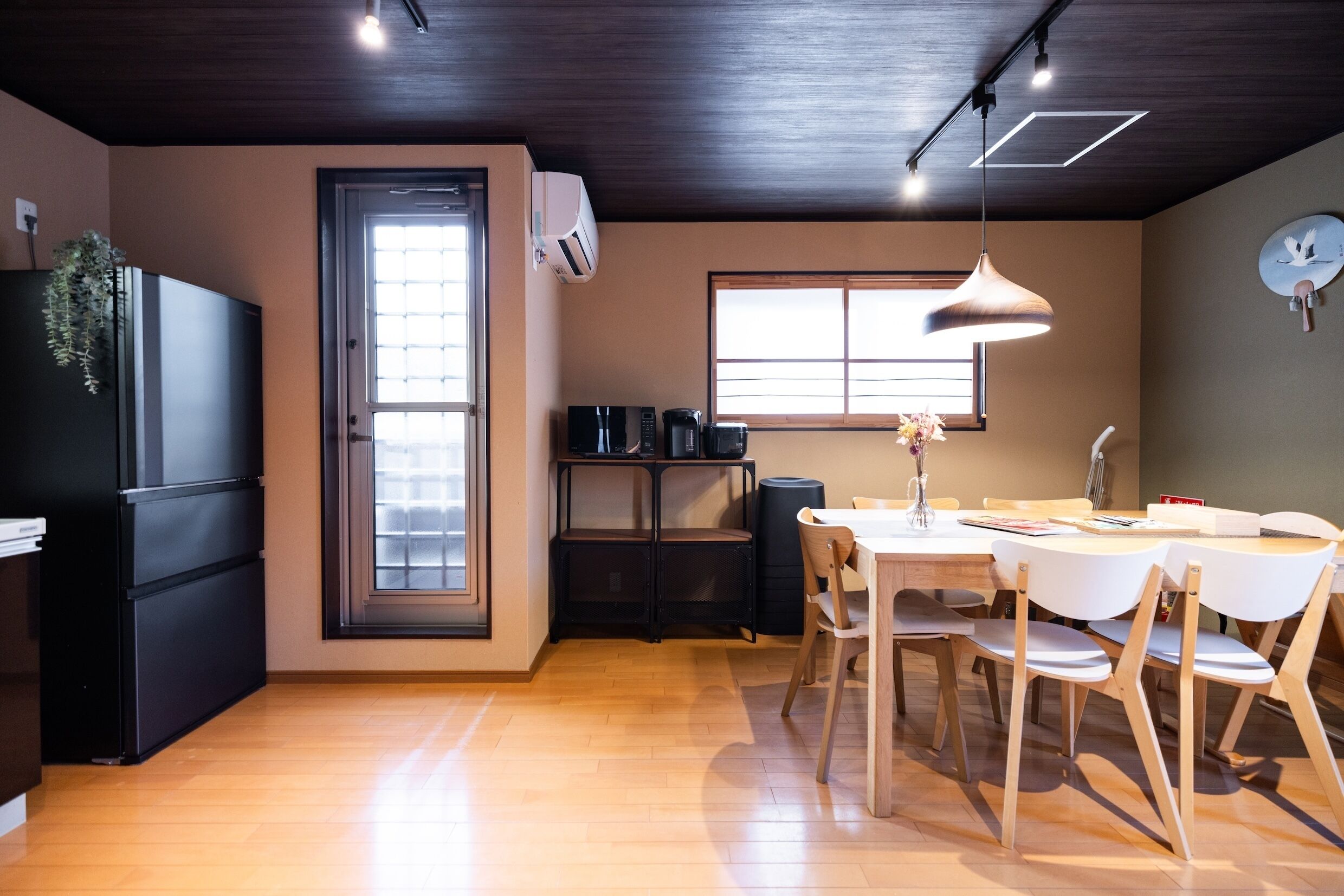 Une cuisine compacte avec un réfrigérateur noir, une table à manger avec des chaises blanches, un luminaire suspendu et une fenêtre avec des stores.