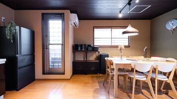 Une cuisine compacte avec un réfrigérateur noir, une table à manger avec des chaises blanches, un luminaire suspendu et une fenêtre avec des stores.