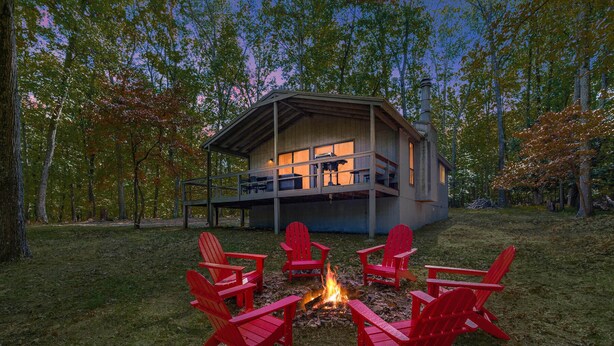 Una cabaña con terraza, rodeada de sillas Adirondack rojas y un brasero.