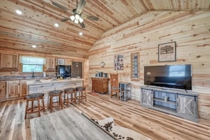 Une cuisine spacieuse aux murs en bois, dotée d’un grand îlot central avec des tabourets, d’un téléviseur encastré et d’un ventilateur de plafond.