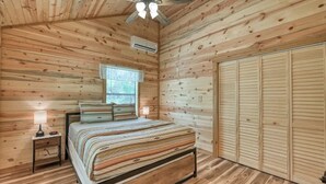 A wooden-paneled bedroom with a bed, bedside lamps, a ceiling fan, and a window. - Benjamin Cabin-HotTub-10 min to Main Street! (Fredericksburg)