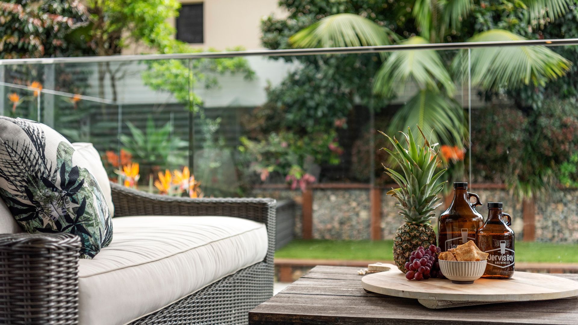 Un sofá de mimbre con un cojín, una mesa redonda de madera con un cuenco de bocadillos, una piña y dos botellas con la etiqueta "Jervis Bay Brewing Co." en un jardín.
