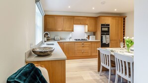 A modern kitchen with wooden cabinets, a central island, and a dining area with white chairs. - Market Square is a cosy, well-equipped holiday home. (Grassington)