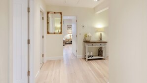 A hallway with a wooden floor, white walls, and a white door on the left. There is a mirror on the wall, a lamp on a table, and a small shelf with decorative items. - Market Square is a cosy, well-equipped holiday home. (Grassington)