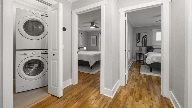 A laundry room with a front-loading washer and dryer, a hallway leading to a bedroom with a bed and a ceiling fan, and a second bedroom with a bed and a nightstand.