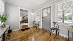 A spacious room with hardwood flooring, a dining area with chairs, a door with a window, and a small table with a plant.