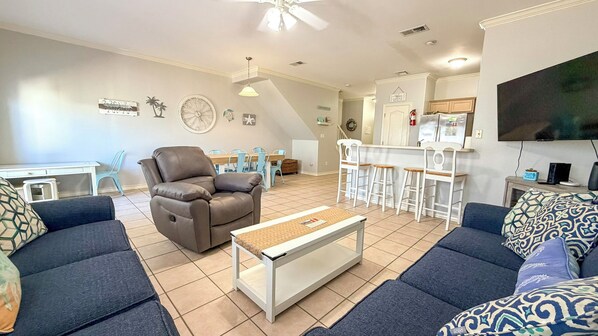 A living room with a brown recliner, two blue sofas, a coffee table, a dining area with blue chairs, a kitchen with a refrigerator, and a TV mounted on the wall.